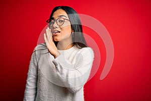 Young beautiful asian woman wearing casual sweater and glasses over red background hand on mouth telling secret rumor, whispering