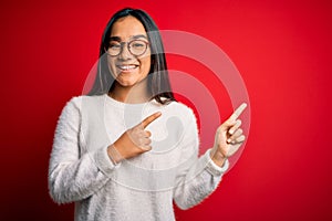 Young beautiful asian woman wearing casual sweater and glasses over red background smiling and looking at the camera pointing with