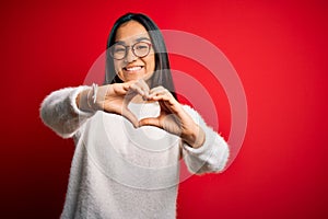Young beautiful asian woman wearing casual sweater and glasses over red background smiling in love doing heart symbol shape with