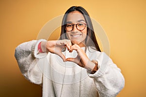 Young beautiful asian woman wearing casual sweater and glasses over yellow background smiling in love doing heart symbol shape