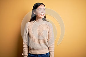 Young beautiful asian woman wearing casual sweater over yellow isolated background looking away to side with smile on face,