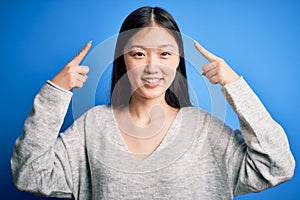 Young beautiful asian woman wearing casual sweater standing over blue isolated background smiling pointing to head with both hands