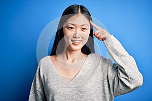 Young beautiful asian woman wearing casual sweater standing over blue isolated background Smiling pointing to head with one