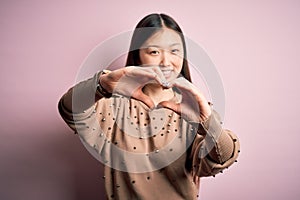 Young beautiful asian woman wearing fashion and elegant sweater over pink solated background smiling in love doing heart symbol