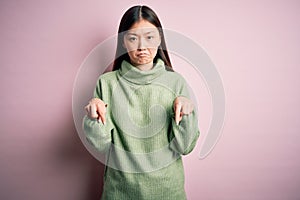 Young beautiful asian woman wearing green winter sweater over pink solated background Pointing down looking sad and upset,