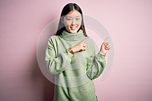 Young beautiful asian woman wearing green winter sweater over pink solated background smiling and looking at the camera pointing