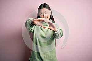 Young beautiful asian woman wearing green winter sweater over pink solated background smiling in love doing heart symbol shape