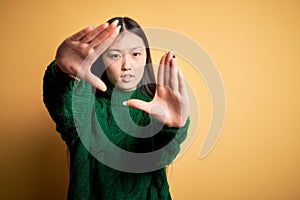 Young beautiful asian woman wearing green winter sweater over yellow  background doing frame using hands palms and