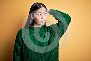 Young beautiful asian woman wearing green winter sweater over yellow isolated background confuse and wondering about question