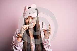 Young beautiful asian woman wearing pajama and sleep mask brushing her teeth using tooth brush, oral paste and glass of water,