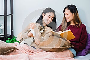 Young beautiful Asian women lesbian couple lover reading book and playing cute cat pet in living room at home with smiling face.