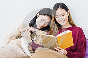 Young beautiful Asian women lesbian couple lover reading book and playing cute cat pet in living room at home with smiling face.
