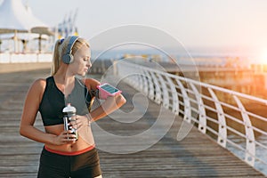 Young beautiful athletic girl with long blond hair in headphones and a bottle of water in hands looks at mobile phone on hand by t