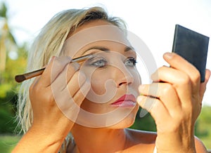 Young beautiful and attractive blond woman with blue eyes retouching makeup with brush applying eyeshadow holding small mirror in