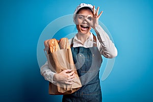 Young beautiful baker woman with blue eyes holding paper bag with  bread with happy face smiling doing ok sign with hand on eye