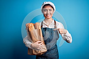 Young beautiful baker woman with blue eyes holding paper bag with  bread with surprise face pointing finger to himself