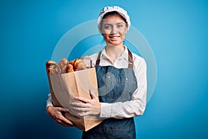 Young beautiful baker woman with blue eyes wearing apron holding paper bag with croissants with a happy face standing and smiling