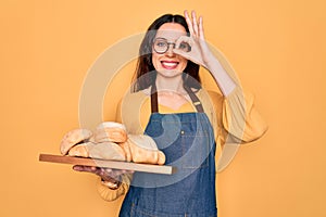 Young beautiful baker woman with blue eyes wearing apron holding tray with bread with happy face smiling doing ok sign with hand
