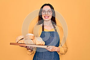 Young beautiful baker woman with blue eyes wearing apron holding tray with bread with a happy face standing and smiling with a