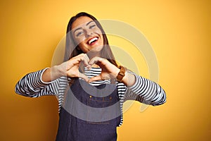 Young beautiful baker woman wearing apron standing over isolated yellow background smiling in love showing heart symbol and shape
