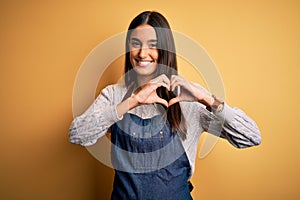 Young beautiful baker woman wearing apron uniform cooking over yellow background smiling in love showing heart symbol and shape