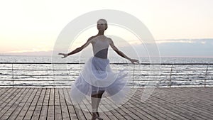 Young, beautiful ballet dancer performing pirouette on embankment near the sea or ocean. Smiling girl in white long tutu