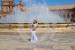 A young and beautiful belly dancer dancing in a square. She is dressed in light blue with a white veil in her hands. World