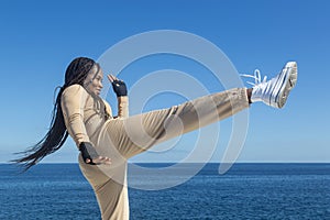 Young beautiful black afro girl training, throwing a kick, blue sky and sea background, attitude and inspiration,