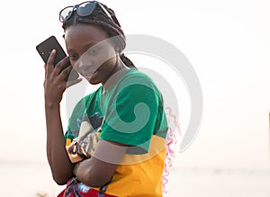 Young beautiful black girl using smart phone on sea beach at sunset