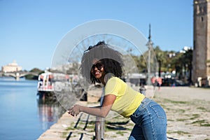 Young and beautiful black woman with afro hair and sunglasses wearing jeans and yellow shirt leaning on the railing overlooking