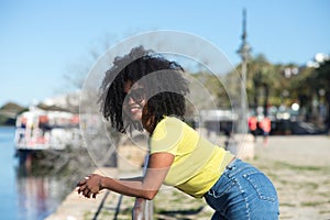 Young and beautiful black woman with afro hair and sunglasses wearing jeans and yellow shirt leaning on the railing overlooking