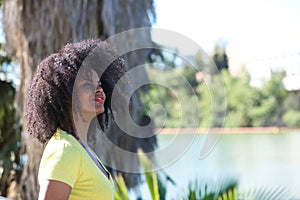 Young, beautiful black woman with afro hair wearing jeans and yellow shirt looking at the river from the shore. The woman is happy