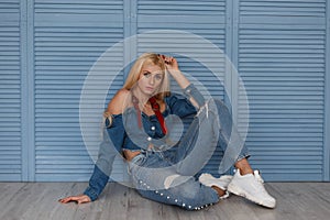 Young beautiful blond girl model in fashion denim clothes with stylish jeans and white sneakers sitting near the blue wooden wall.