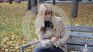 Young beautiful blond girl sitting on a bench in an autumn park with a phone in hands