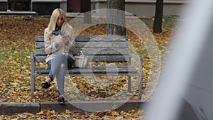 Young beautiful blond girl sitting on a bench in an autumn park with a phone in hands