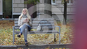 Young beautiful blond girl sitting on a bench in an autumn park with a phone in hands