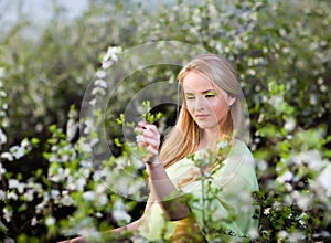 Young beautiful blond smiling woman in yellow dress standing in blooming cherry trees and looking at flower