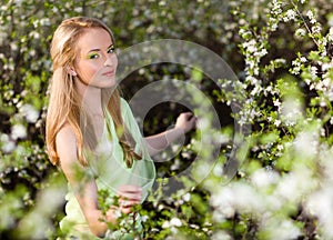 Young beautiful blond smiling woman in yellow dress standing in blooming cherry trees and looking at flower