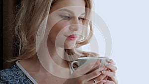 A young beautiful blond woman in a blue robe stands by the window of a room and drinks tea or coffee from a white cup.