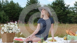 A young beautiful blond woman in a hat and dress is drinking lemonade from a can while sitting on a plaid on the green