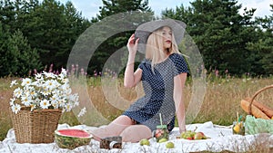 A young beautiful blond woman in a hat and dress is drinking lemonade from a can while sitting on a plaid on the green