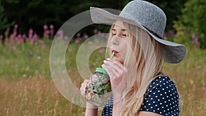 A young beautiful blond woman in a hat and dress is drinking lemonade from a can while sitting on a plaid on the green