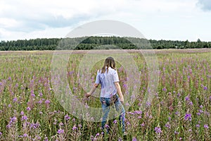 Young beautiful blond woman in purple shirt from behind walking in the meadow among flowers of fireweed