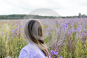 Young beautiful blond woman in purple shirt from behind walking in the meadow among flowers of fireweed