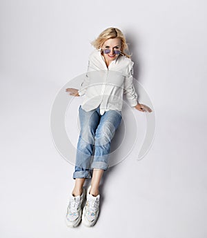 Young beautiful blond woman in stylish casual clothing, white sneakers and sunglasses sitting on floor and smiling, top view
