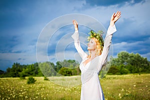 Young beautiful blond woman in white dress and wreath standing with eyes closed and enjoying sunshine on summer day