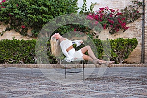 Young, beautiful, blonde American woman in a white dress, with her head back and her hair falling down sitting on a chair in the