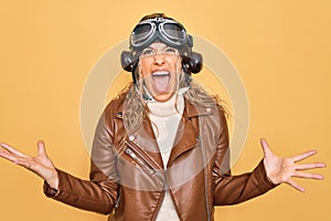 Young beautiful blonde aviator woman wearing vintage pilot helmet whit glasses and jacket crazy and mad shouting and yelling with