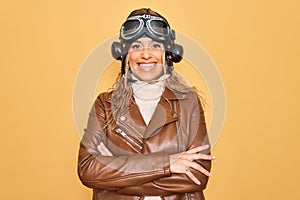 Young beautiful blonde aviator woman wearing vintage pilot helmet whit glasses and jacket happy face smiling with crossed arms