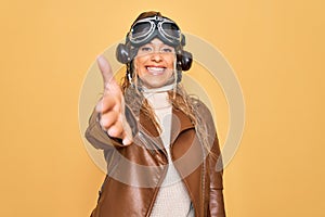 Young beautiful blonde aviator woman wearing vintage pilot helmet whit glasses and jacket smiling friendly offering handshake as